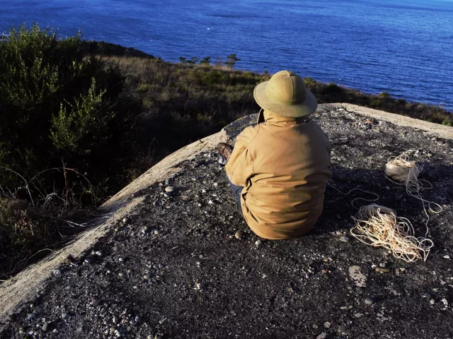 A man sitting with his back to the camera on a bunker overlooking the ocean